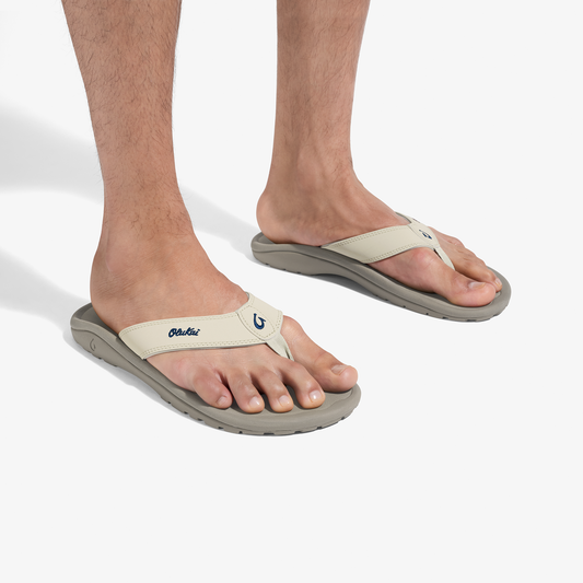 Man wearing cream and gray thong sandals with curved sole and embroidered logo on upper strap.