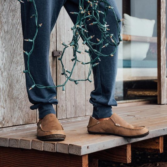 Toffee-colored suede slipper with stitched seams worn with dark jeans on wooden deck.