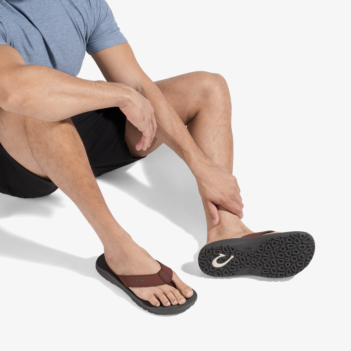 Man wearing blue shirt and black shorts seated while wearing brown and black thong sandal with textured sole.