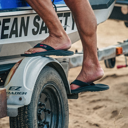 Black flip-flops with pink accents worn on feet resting on a vehicle tire.