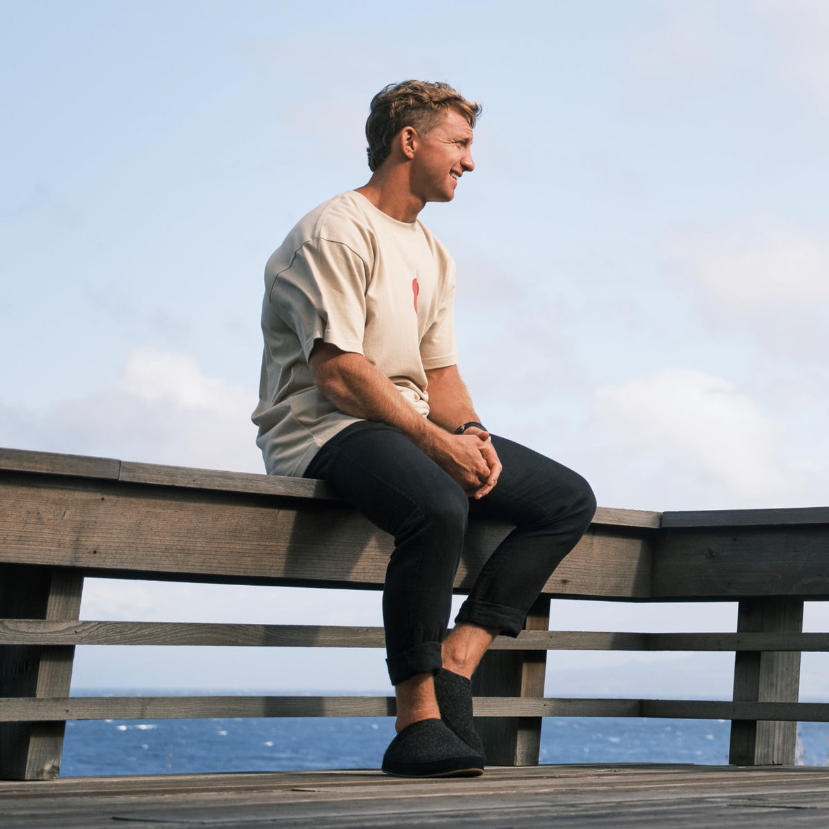 Man wearing beige short-sleeve t-shirt with small red emblem and black pants, sitting on wooden railing.