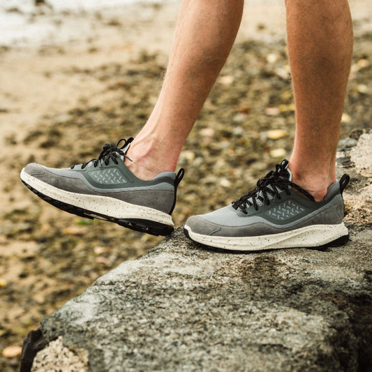 Person wearing grey and white lace-up running shoes with black accents on rocky beach terrain.