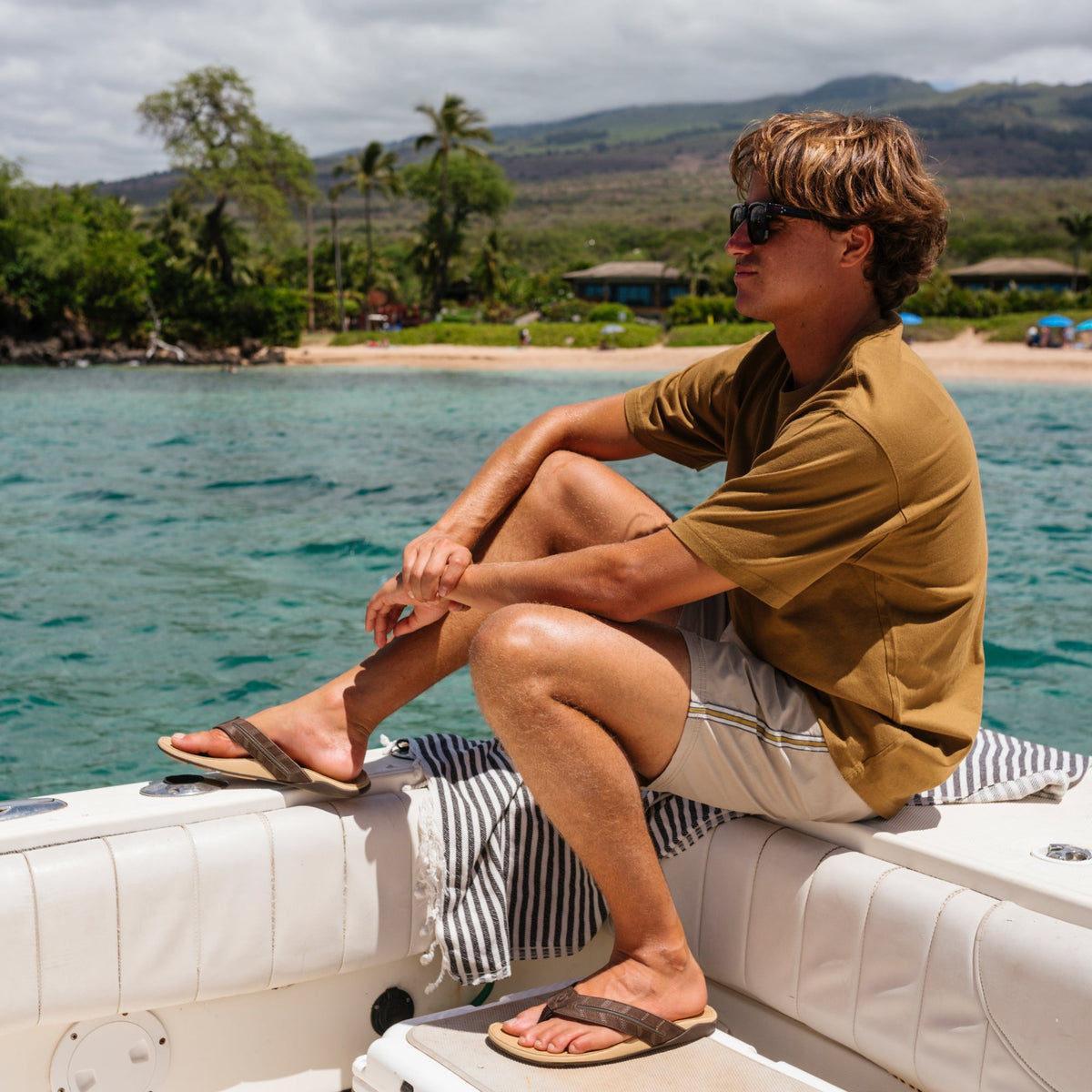 Man wearing golden sand colored short-sleeve shirt and dark navy striped swim trunks, seated on a boat.