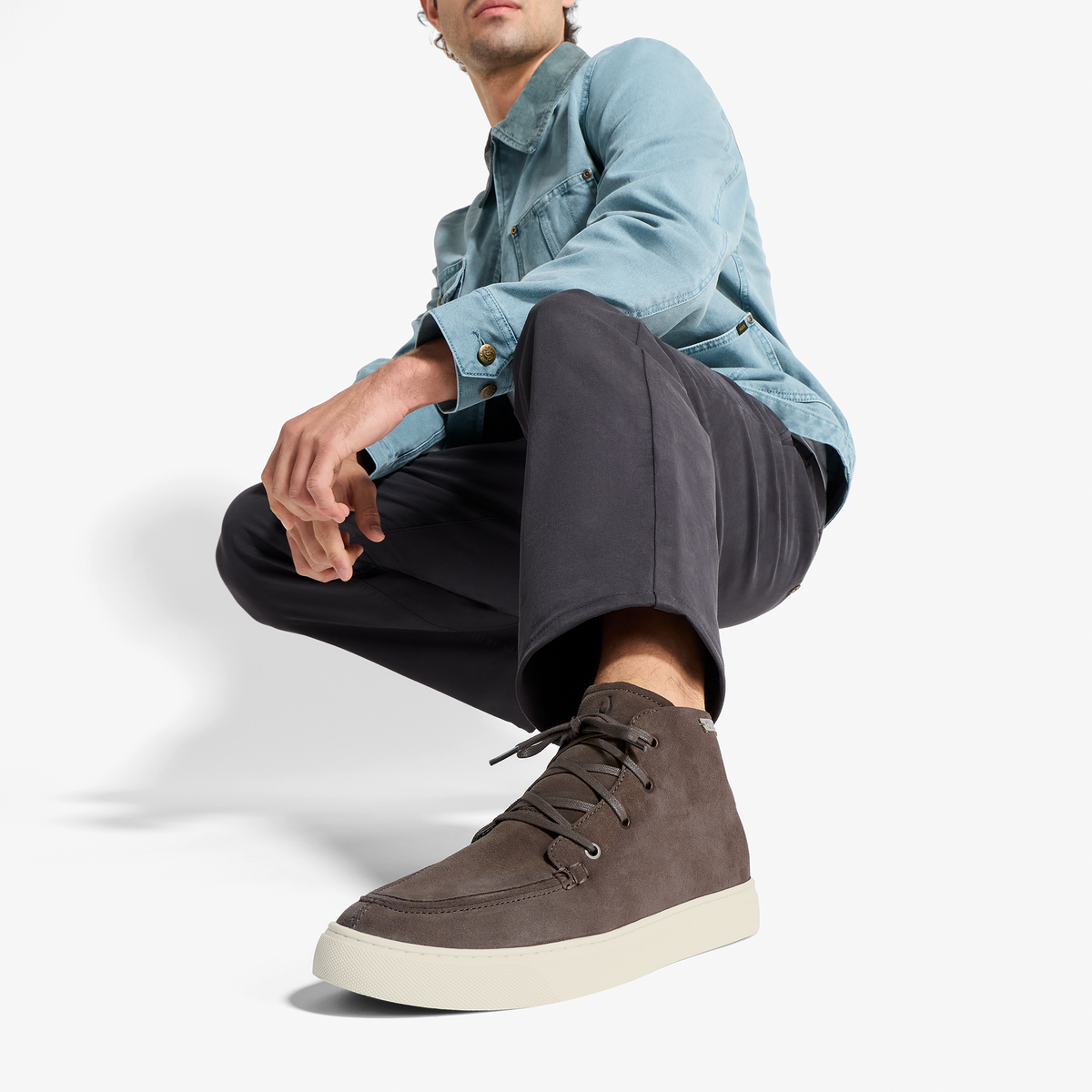 Dark brown suede chukka boot with cream sole, laced closure, worn with black pants and light blue shirt.