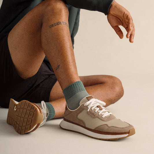 Ecru and taupe grey sneaker with white laces worn with ribbed socks, shown from side angle with visible sole.