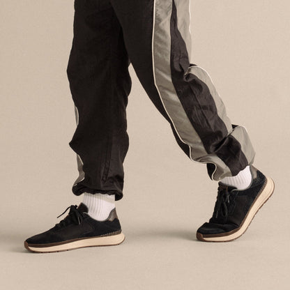 Black suede sneakers with cream sole and laces, worn with black pants with gray side stripe and white socks.