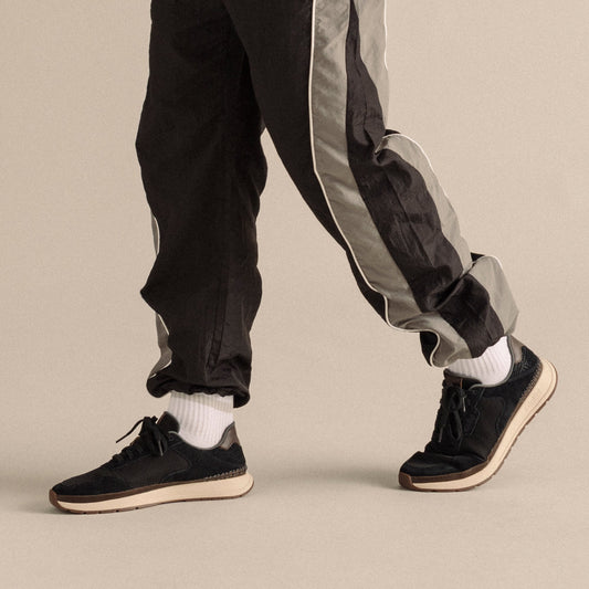 Black suede sneakers with cream sole and laces, worn with black pants with gray side stripe and white socks.