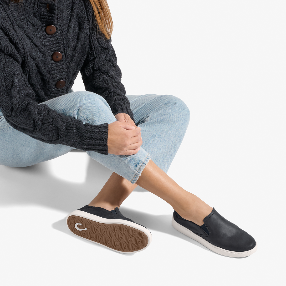 Black leather slip-on sneaker with white sole, worn with light blue jeans and dark cardigan.