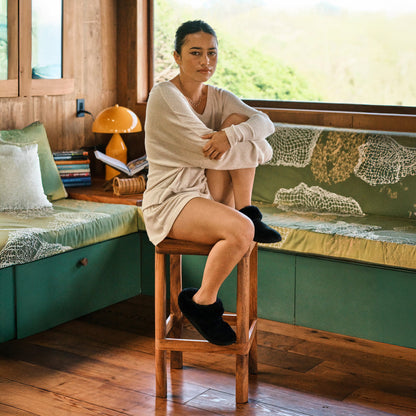 Woman sitting on wooden stool wearing cream-colored ribbed cardigan and black fuzzy slippers in bedroom with green upholstere