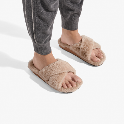 Tan fluffy slide slippers with cushioned footbeds, worn with gray knit pants.