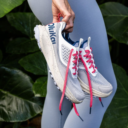 White and coastal blue running shoe with pink laces held in hand against green foliage.