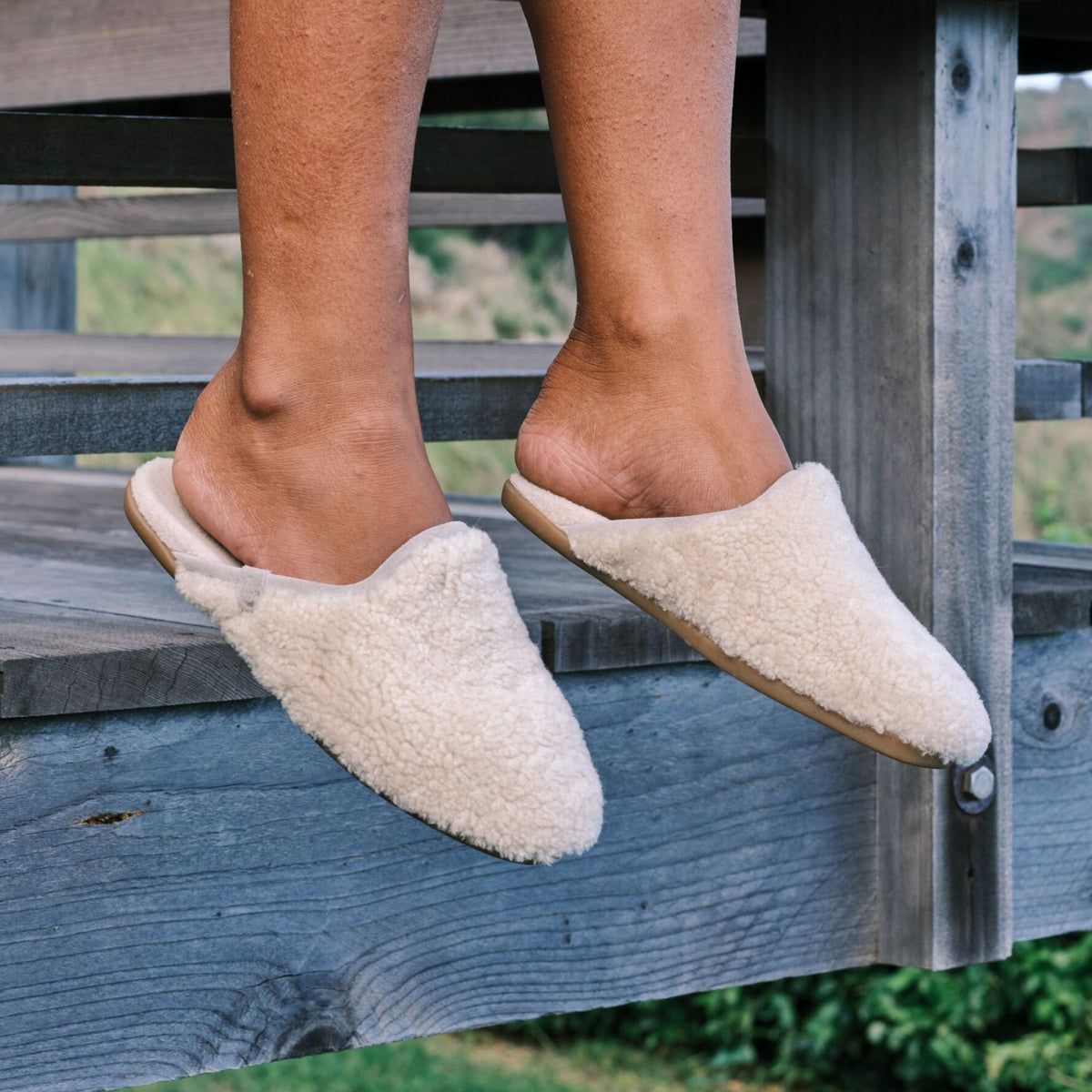 Cream wool mule slippers with tan sole worn by person sitting on wooden bench.