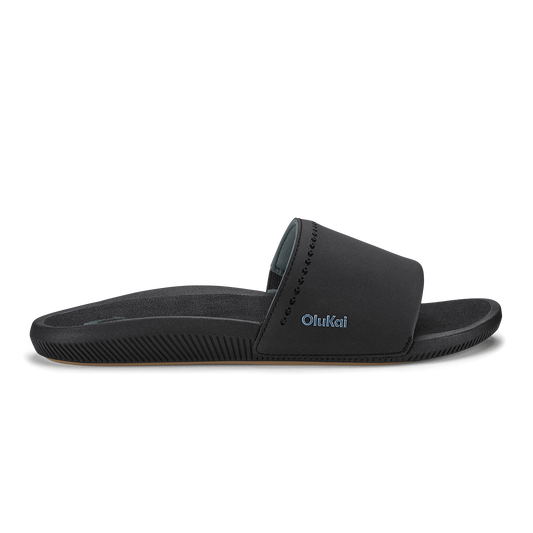 Black slide sandal with padded upper and textured sole featuring OluKai branding.
