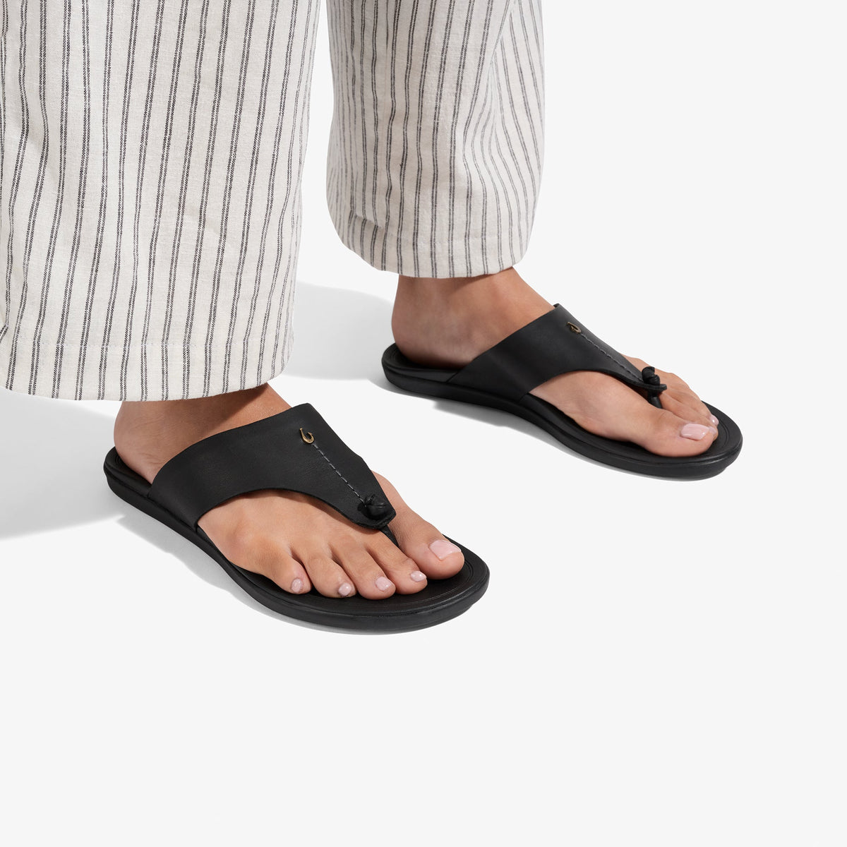 Black thong sandals with padded straps and contoured soles, worn with striped linen pants.