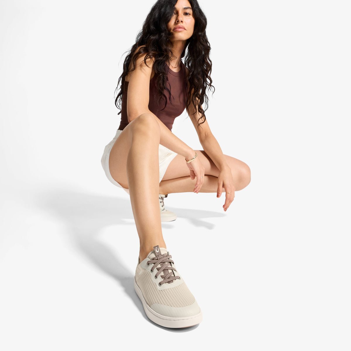 Woman wearing burgundy sleeveless top, cream shorts, and white lace-up sneakers, crouching against white background.