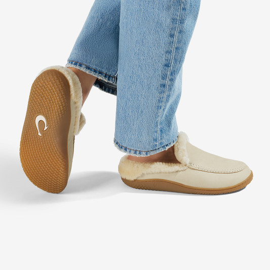 Ecru suede slip-on loafer with sherpa lining and tan rubber sole, shown worn with blue jeans.