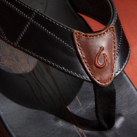 Black sandal with brown leather strap detail and embroidered hook symbol on toe post.